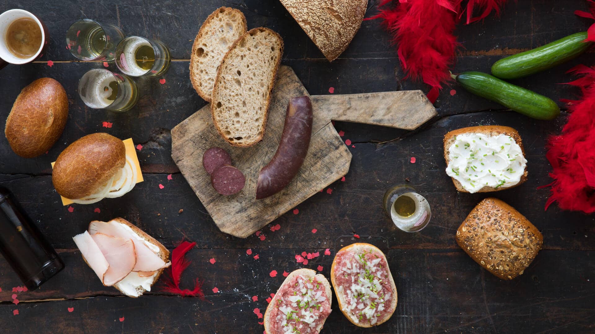 Katerfrühstück. Tisch mit Schwarzwälder Kruste Brot, Blutwurst, Mettbrötchen, Halten Hahn, Kölsch, Espresso, Mehrkornbrötchen mit Frischkäse, Fitnessbrötchen mit Putenbrust, Gurke, Konfetti, Federboa, leere Bierflasche